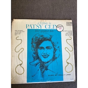 May include: A vintage vinyl record album titled "a Tribute to Patsy Cline: Her Greatest Hits." The album cover features a blue background with a portrait of a woman and the text "Sung by Holly Lane."
