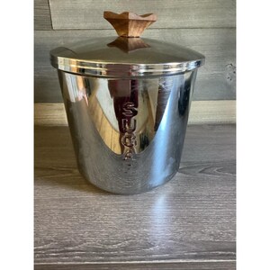 Puede incluir: Un recipiente para azúcar cilíndrico de acero inoxidable con un acabado pulido y reflectante. La palabra SUGAR está grabada verticalmente en la parte frontal. Incluye una tapa a juego con un pomo de madera decorativo en forma de estrella.