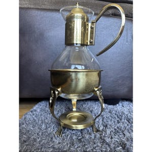 May include: A vintage brass and glass coffee maker with a clear glass carafe and a brass stand. The stand has ornate legs and a circular base. The coffee maker has a brass handle and a lid. The glass carafe has a bulbous shape.