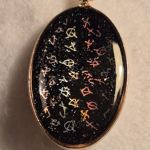 Può includere: Una collana con pendente ovale con catena color oro. Il pendente ha uno sfondo nero con glitter sparsi e simboli colorati. Il pendente è incorniciato con un metallo color oro.