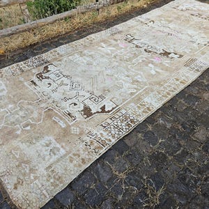 May include: A rectangular, beige rug with an intricate, distressed pattern. The rug features a central design with geometric shapes and floral motifs in shades of brown and white. The rug is placed on a dark, textured surface.