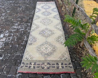 May include: A rectangular, beige rug with a repeating geometric pattern in gray and blue. The rug has a red fringe on the bottom edge. The rug is displayed on a cobblestone surface.