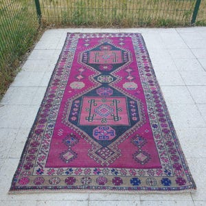 Puede incluir: Una alfombra rectangular vintage con un vibrante patrón geométrico rosa y azul marino. La alfombra presenta un diseño central con formas de diamantes y cuadrados, rodeado de intrincados bordes en colores complementarios. La alfombra está colocada sobre una superficie de baldosas de color claro.