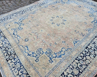 May include: Large, rectangular area rug with a central floral design in shades of beige and pink. The rug features a wide border with intricate blue floral patterns and a dark blue outer edge. The rug is placed on a stone surface.