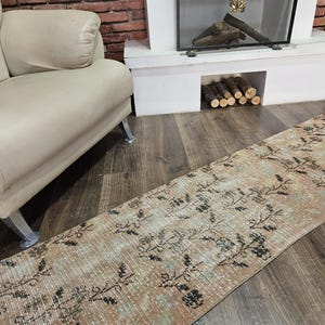 May include: A rectangular, distressed-look rug with a floral pattern in shades of beige, brown, and black. The rug is placed on a wooden floor near a beige sofa and a white fireplace.