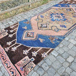 May include: A rectangular rug with a central design in tan, blue, and pink. The rug features geometric patterns and borders in shades of brown, blue, and pink. The rug is placed on a cobblestone surface.