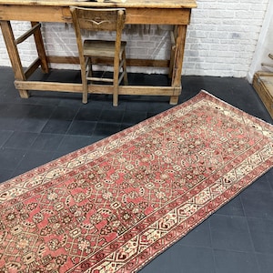 Puede incluir: Una alfombra larga y rectangular, vintage, con un estampado floral rojo y crema. La alfombra tiene un borde detallado y está colocada sobre un suelo de baldosas oscuras. La alfombra es un revestimiento decorativo para el suelo.