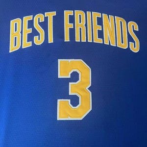 May include: A blue athletic jersey featuring the text BEST FRIENDS in yellow block letters with white outlines, and the number 3 in yellow with a white outline below. The neckline has a white and yellow striped ribbed trim design.