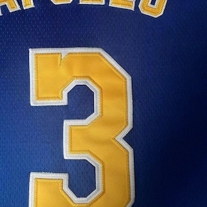 May include: A royal blue athletic jersey featuring the name APOLLO in yellow block letters with white trim, positioned above a large yellow number 3 with white trim. The fabric has a visible mesh texture throughout the entire garment surface.