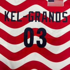 以下が含まれることがあります： 赤と白の波状ストライプ柄のジャージで、濃い青色の文字で「KEL-GRANDS」と書かれ、同じく濃い青色で数字の「03」がプリントされています。前面の右上隅には小さなアメリカ国旗のグラフィックが配置されています。