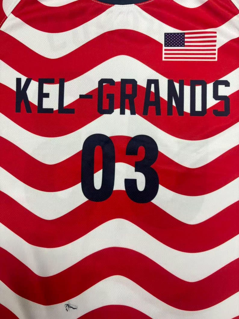 以下が含まれることがあります： 赤と白の波状ストライプ柄のジャージで、濃い青色の文字で「KEL-GRANDS」と書かれ、同じく濃い青色で数字の「03」がプリントされています。前面の右上隅には小さなアメリカ国旗のグラフィックが配置されています。