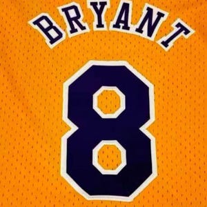 May include: A gold basketball jersey featuring the name BRYANT arched in dark purple letters with a white outline. Below the name is the large number 8 in dark purple with a white border. The fabric has a distinct mesh texture throughout the garment.