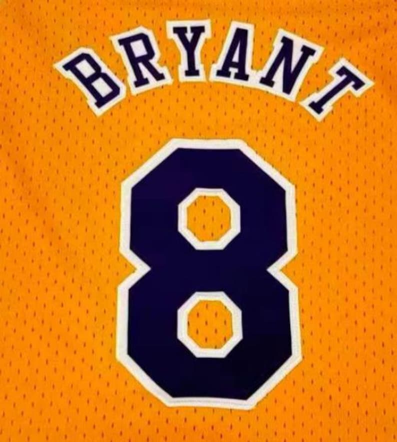May include: A gold basketball jersey featuring the name BRYANT arched in dark purple letters with a white outline. Below the name is the large number 8 in dark purple with a white border. The fabric has a distinct mesh texture throughout the garment.