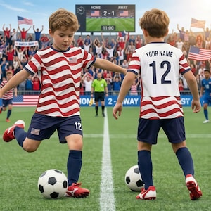 May include: Two young soccer players in red, white, and blue striped jerseys and navy shorts are on a green field. One kicks a soccer ball. The scoreboard shows the score as 2-1. The jerseys have the number 12.