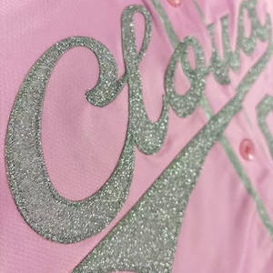 May include: A light pink button-down shirt featuring the word Cloud in large, silver glitter script lettering across the front. The garment includes matching pink buttons and a silver glitter tail design extending from the text.