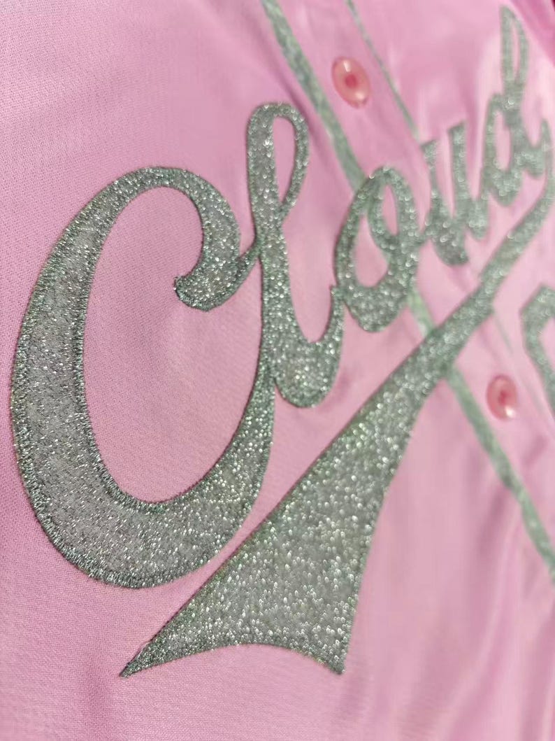 May include: A light pink button-down shirt featuring the word Cloud in large, silver glitter script lettering across the front. The garment includes matching pink buttons and a silver glitter tail design extending from the text.