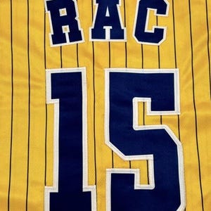 May include: A yellow athletic jersey featuring thin vertical black pinstripes. The back displays the letters RAC in dark blue with white trim, positioned above the large number 15, also rendered in dark blue with white trim.