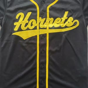 May include: A black baseball jersey featuring a button-down front with yellow piping along the placket and collar. The word "Hornets" is embroidered in yellow script across the chest, with a yellow underline extending from the letter s.