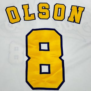 May include: A white sports jersey featuring the name OLSON in bold yellow block letters with a dark blue outline, positioned above the large yellow number 8, which also features a dark blue outline, set against a white mesh fabric background.