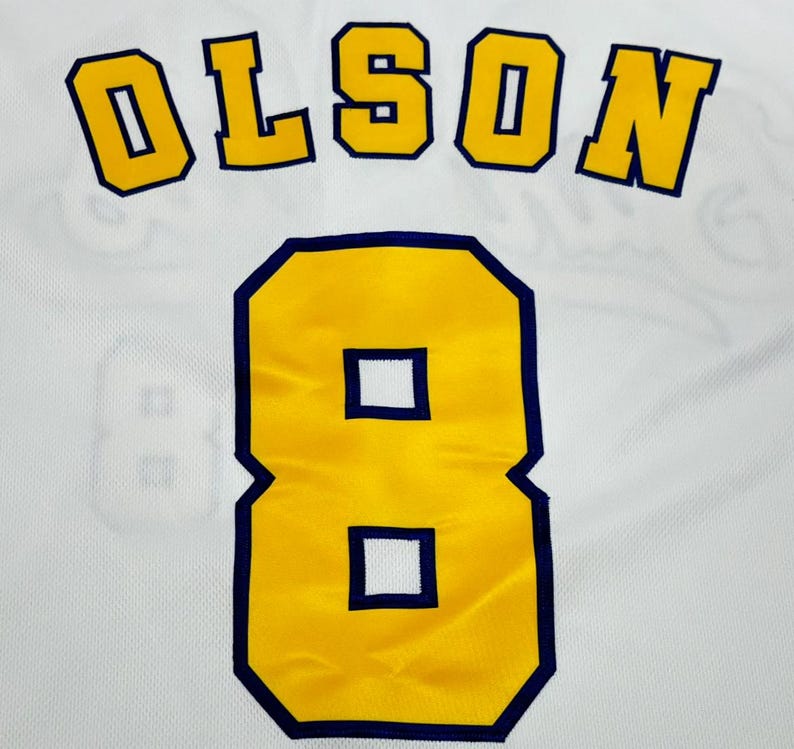 May include: A white sports jersey featuring the name OLSON in bold yellow block letters with a dark blue outline, positioned above the large yellow number 8, which also features a dark blue outline, set against a white mesh fabric background.