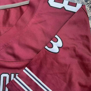 Puede incluir: Camiseta deportiva granate con detalles en blanco y azul marino. La camiseta lleva el n&uacute;mero "3" en blanco con contorno negro en la manga y el hombro. La camiseta tiene cuello en V y una raya blanca en el cuello.