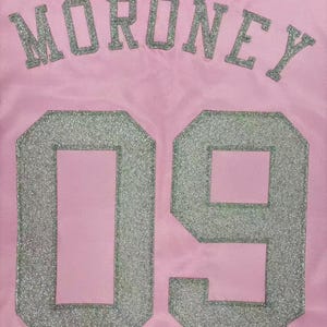 May include: A light pink jersey featuring the name MORONEY in silver glitter lettering arched above the large silver glitter number 09. The fabric has a subtle mesh texture, and the lettering and numbers are applied with a distinct glitter finish.