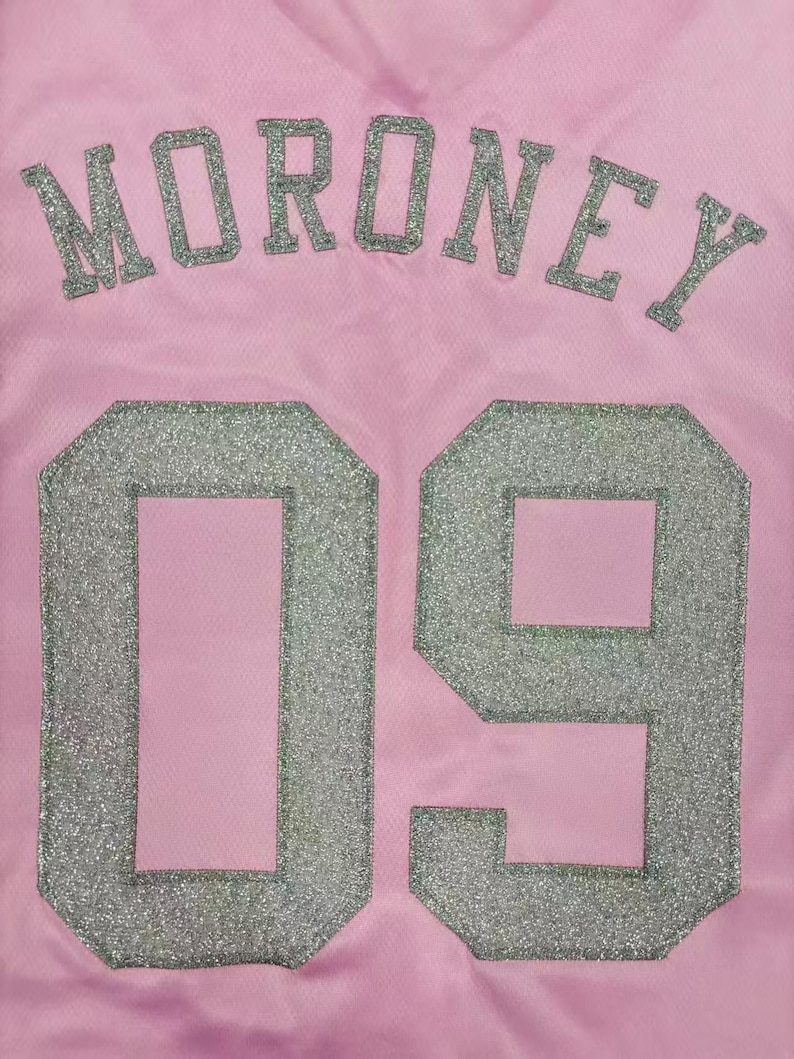 May include: A light pink jersey featuring the name MORONEY in silver glitter lettering arched above the large silver glitter number 09. The fabric has a subtle mesh texture, and the lettering and numbers are applied with a distinct glitter finish.