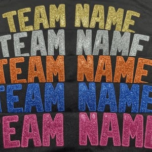 May include: A black fabric surface featuring five rows of the text "TEAM NAME" in glittery heat-applied lettering. The rows appear in gold, silver, orange, blue, and pink, each displaying a textured, sparkling finish against the dark background.
