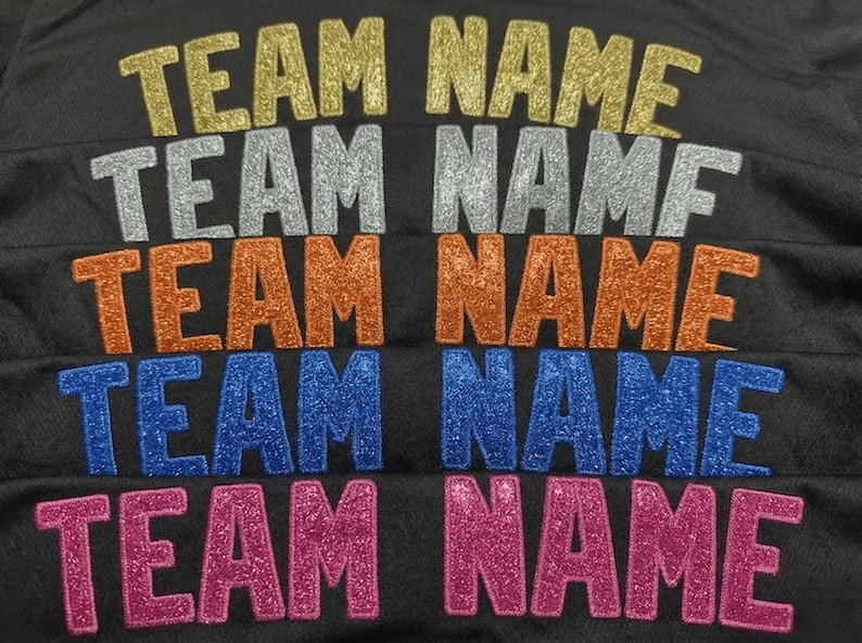 May include: A black fabric surface featuring five rows of the text "TEAM NAME" in glittery heat-applied lettering. The rows appear in gold, silver, orange, blue, and pink, each displaying a textured, sparkling finish against the dark background.