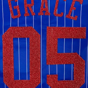 May include: A royal blue fabric with thin vertical white pinstripes. The name GRACE is appliqued in red glitter block letters above the large red glitter numbers 05. The texture of the red lettering is coarse and sparkling against the smooth blue.
