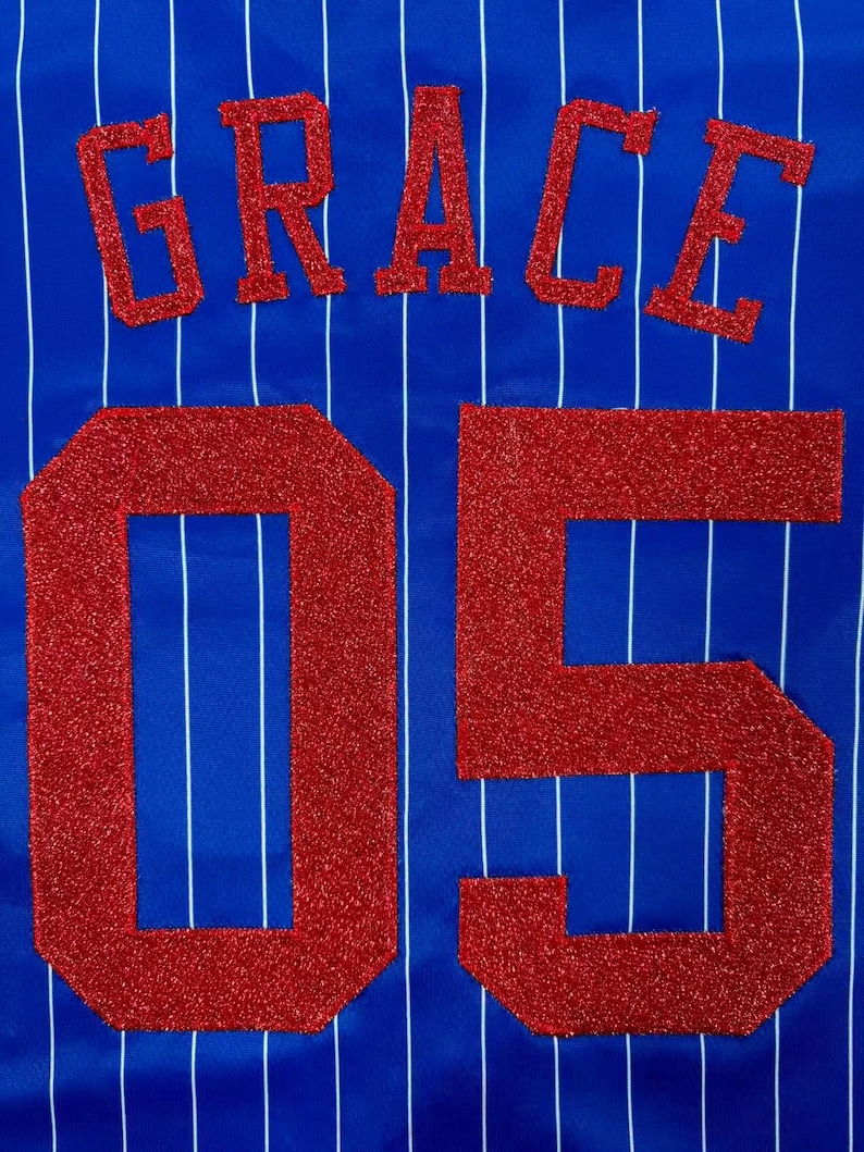 May include: A royal blue fabric with thin vertical white pinstripes. The name GRACE is appliqued in red glitter block letters above the large red glitter numbers 05. The texture of the red lettering is coarse and sparkling against the smooth blue.