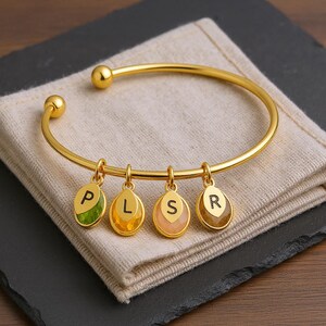 Puede incluir: Una pulsera dorada con cuatro colgantes. Cada colgante tiene un color diferente y una letra: P, L, S y R. La pulsera descansa sobre una tela beige, colocada sobre una superficie gris oscuro.