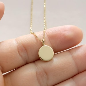 May include: A gold-colored necklace with a small, round pendant. The pendant is smooth and shiny, and the chain is delicate. The necklace is held in a hand, suggesting it is being displayed or worn. The jewelry is simple and elegant.