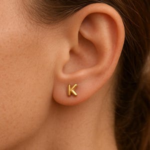 Może przedstawiać: Złota kolczyka w kształcie litery "K" jest widoczna na uchu. Kolczyk jest mały i prosty, z wypolerowanym wykończeniem. Kolczyk to klasyczna i wszechstronna biżuteria.