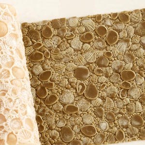 Puede incluir: Un rodillo cilíndrico de color beige con un patrón junto a una hoja cuadrada de material color canela. Ambos artículos presentan una textura en relieve detallada y repetitiva de varias conchas marinas pequeñas.