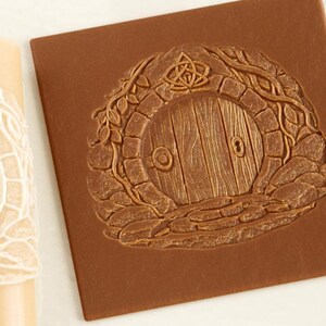 May include: A cylindrical beige rolling pin with a recessed circular door design and a square brown clay slab featuring the same embossed door motif, which includes a stone archway, a wooden door, and a small decorative knot symbol above.