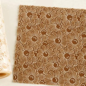Könnte beinhalten: Ein zylindrisches Nudelholz mit erhabenem Gänseblümchenmuster und ein quadratisches Blatt aus braunem Material mit demselben geprägten Blumenmuster. Das Nudelholz ist hellbeige, während das quadratische Blatt eine dunkelbraune Blumenstruktur zeigt.