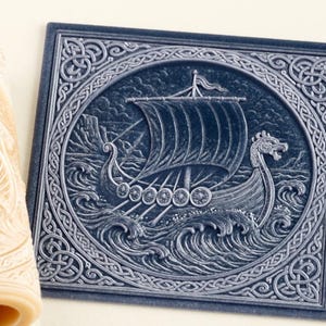May include: A cylindrical beige rolling pin and a square dark blue tile, both featuring a relief carving of a Viking longship sailing on waves. The ship has a large sail and a dragon-headed prow, surrounded by an intricate Celtic knot border.