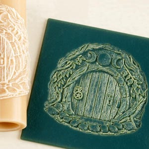 May include: A cylindrical beige rolling pin with a carved relief pattern of a round door surrounded by foliage and a triple moon symbol, alongside a square dark green tile featuring the same embossed door design pressed into its surface.