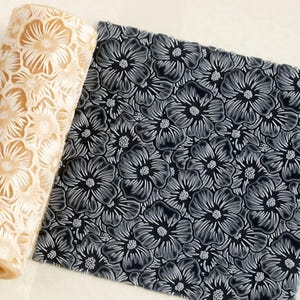 Può includere: Un mattarello cilindrico con un motivo floreale intagliato e un foglio quadrato di materiale grigio scuro con un disegno floreale in rilievo che corrisponde al motivo sul mattarello, mostrando la texture creata dallo strumento.