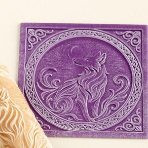 May include: A cylindrical tan rolling pin featuring an embossed wolf design and a square purple tile with a matching embossed wolf motif inside a circular border with intricate scrollwork patterns in the corners of the square frame.