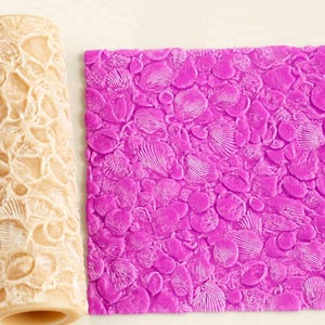 May include: A cylindrical beige rolling pin with a carved seashell pattern next to a square sheet of bright purple material featuring an embossed, repeating texture of various seashells, including scallops and spiral shells, across the surface.