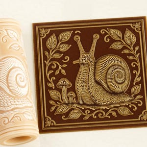 Pode incluir: Um rolo de massa cilíndrico bege e um azulejo quadrado castanho escuro, ambos com um design em relevo de um caracol rodeado por folhas e pequenos cogumelos. O padrão do caracol repete-se no rolo e está centrado no azulejo.