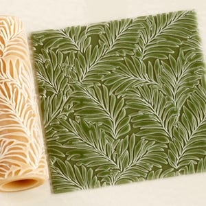 May include: A cylindrical beige rolling pin with a raised fern leaf pattern next to a square piece of green clay that has been impressed with the same leaf design, showing the texture created by the tool on the surface of the material.