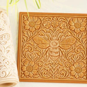 May include: A cylindrical rolling pin and a square flat stamp, both featuring a deeply embossed pattern of a central bee surrounded by intricate floral and leafy scrollwork. The rolling pin is light beige, while the square stamp is tan.