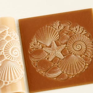 Puede incluir: Un rodillo cilíndrico de color beige con un patrón en relieve de conchas marinas, estrellas de mar y coral, junto a una baldosa de arcilla cuadrada de color marrón que muestra el mismo diseño intrincado de conchas y estrellas de mar.