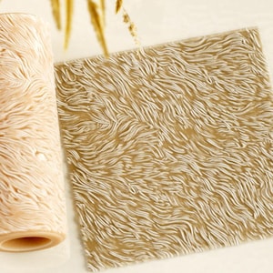 Op de afbeelding: Een cilindrische patroonverfroller en een vierkant plat vel met een verhoogde, wervelende, haarachtige textuur in een lichtbeige kleur. De roller heeft een hol midden en het oppervlak van beide items toont een consistent, vloeiend, organisch patroon.