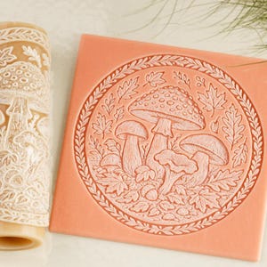 May include: A cylindrical beige rolling pin and a square peach-colored flat stamp, both featuring an embossed design of various mushrooms, oak leaves, and acorns arranged within a circular border of delicate foliage and vines.