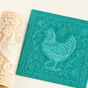 Puede incluir: Un rodillo cilíndrico de color beige y una lámina cuadrada de color verde azulado, ambos con un diseño en relieve de una gallina rodeada de enredaderas florales y un borde geométrico. Los patrones en relieve son idénticos en ambos objetos.