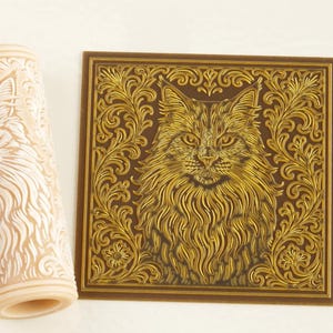 May include: A cylindrical beige rolling pin with a deeply carved cat and scrollwork pattern alongside a square brown tile featuring the same cat and scrollwork design in raised gold relief, showcasing the embossed texture of the cat's fur.